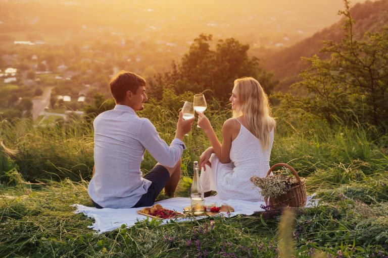 Loving young couple drinking wine at sunset on the hill. A boyfriend and a girl on a romantic date, sitting and clinking glasses with wine.